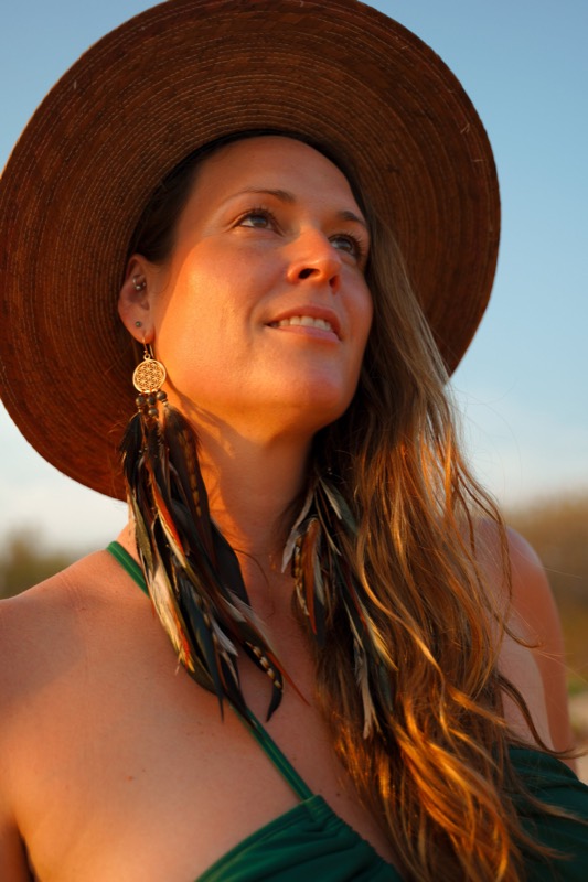 A woman in a wide-brimmed hat at golden hour wearing long brown, green and light feather earrings with a gold Flower of Life charm