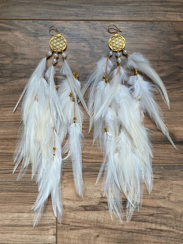 White feather earrings with gold Flower of Life charms laid on a wooden table